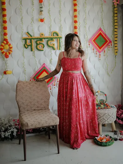 A full-length portrait of the bride at her Mehndi ceremony. She stands beside the festive decor, her bright pink outfit and joyful expression lighting up the frame.