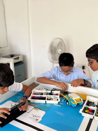 A team of three students huddles around their robot and a tablet, preparing for the IRO Techies category. They are using block-based coding to program their robot's movements and actions.