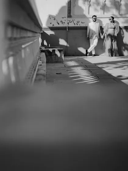An artistic black and white shot using a low angle and foreground blur. This creative composition creates a sense of scale and drama, focusing on the couple against the wall.