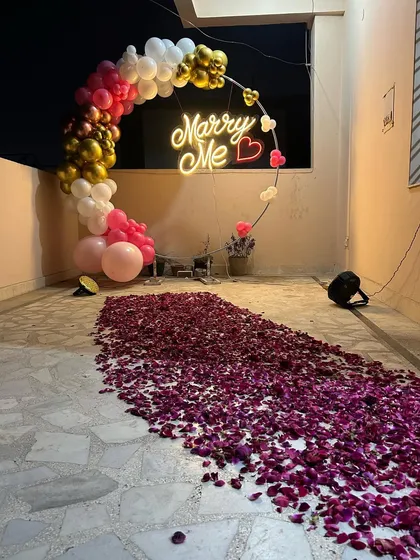 A modern proposal setup on a terrace, combining a circular balloon arch with a 'Marry Me' neon sign. A path of rich purple flower petals leads the way, creating a colorful and contemporary romantic scene.