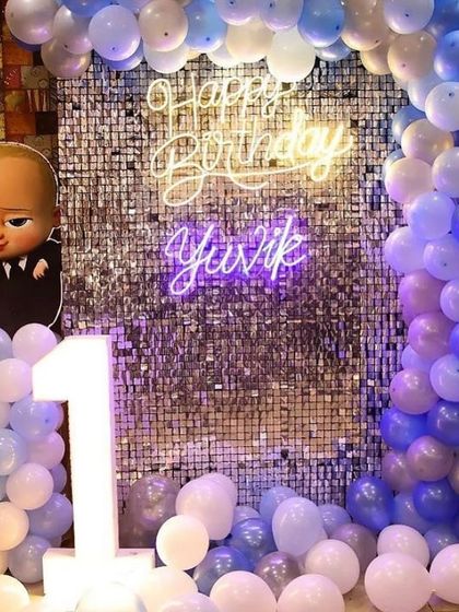 A vibrant Boss Baby themed first birthday party. The decor features a silver shimmer wall, a blue and white balloon arch, and a 'Happy Birthday' neon sign.