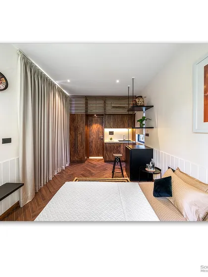 The interior of a compact prefab home, showcasing a clever use of space with a combined bedroom and kitchenette. The warm wood tones and large window create a cozy and functional living environment.