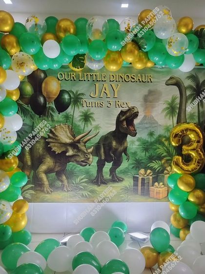 A thrilling dinosaur theme party for a 3-year-old, featuring a large T-Rex backdrop and a green and gold balloon arch.