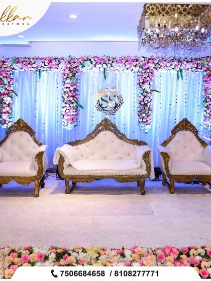 A royal reception stage with a white and gold theme. The ornate sofas and backdrop of white drapes and purple flowers create a look of pure elegance.