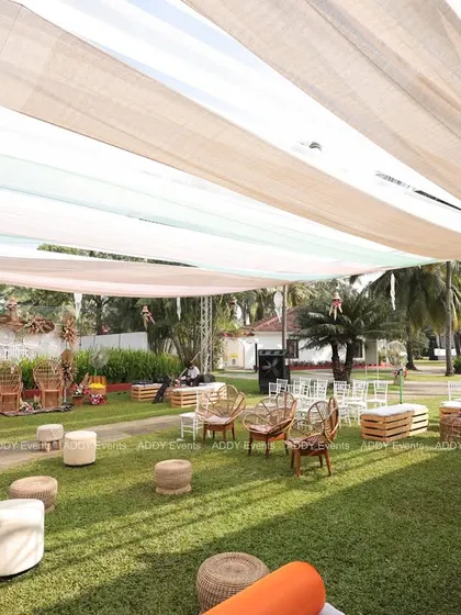 A wide view of a relaxed, boho-themed Mehndi seating area on a lush lawn, with scattered wicker chairs, poufs, and a light fabric canopy overhead.