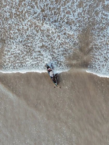 A top-down drone shot of a boat meeting the surf. The contrast between the calm sand and the frothy white waves creates a beautiful, minimalist composition.
