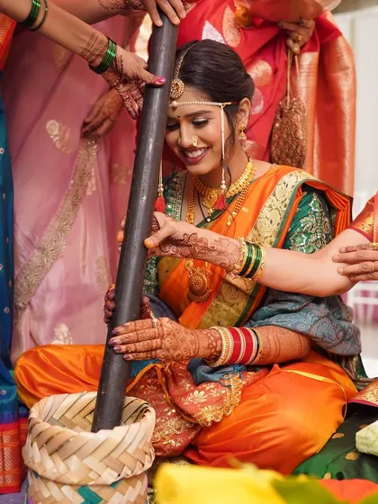 Capturing the beauty of tradition. The bride participates in a wedding ritual, her smile lighting up the frame. The orange and green saree is a classic choice for Maharashtrian ceremonies.