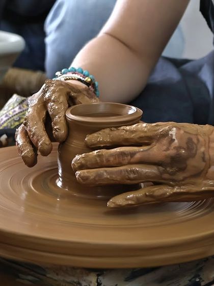 A close-up of hands covered in rich, brown clay, gently guiding a small cup into shape on the potter's wheel. It's all about the connection.