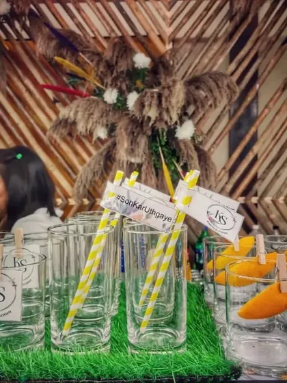A rustic and chic bar setup detail. Glasses are prepped with personalized initial tags and yellow-striped straws, ready for service against a backdrop of woven pampas grass decor.