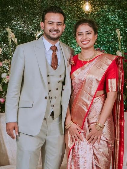 The happy couple, Ivy and Vivek, posing for a photo. The groom's light grey suit and the bride's red and gold saree look stunning against the modern, nature-inspired decor.