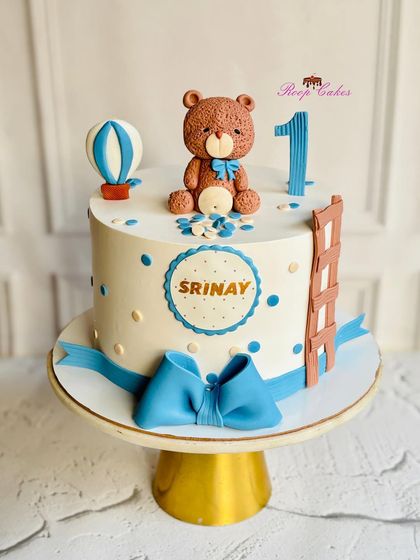 A sweet teddy bear themed cake for Srinay's first birthday. This fondant cake features a cute bear, a hot air balloon, and a big blue bow, creating a classic and adorable look.