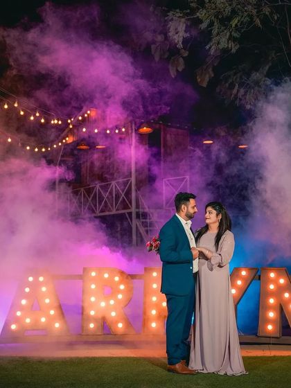 A wide shot showing the full scale of our 'MARRY ME' light-up sign setup, with a couple standing amidst colorful smoke. This demonstrates the grand and theatrical scenes we can create for your pre-wedding shoot.