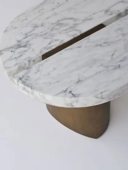A close-up of the SPLIT center table, which features a solid Statuario marble top elegantly divided by a metal accent. The base is crafted from cast metal, providing a sculptural foundation.