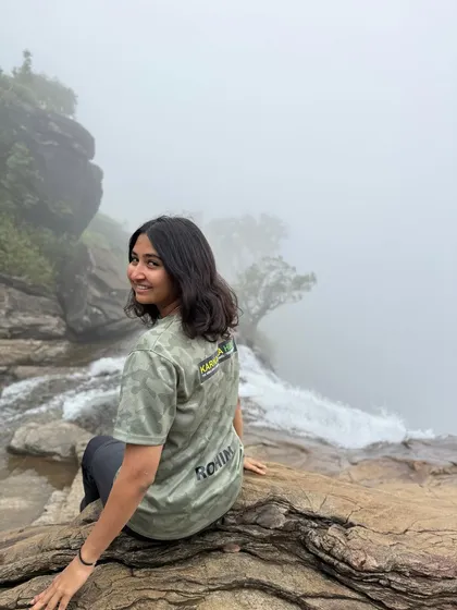 A trekker wearing our Karnataka Hikes t-shirt sits at the edge of Bandaje Falls, looking back with a smile.