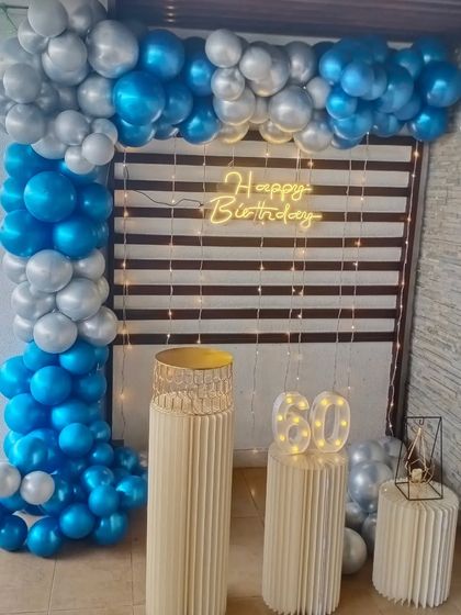 An elegant backdrop for a 60th birthday. This setup features a blue and silver balloon arch against a window blind backdrop, with light-up marquee numbers and pedestals.