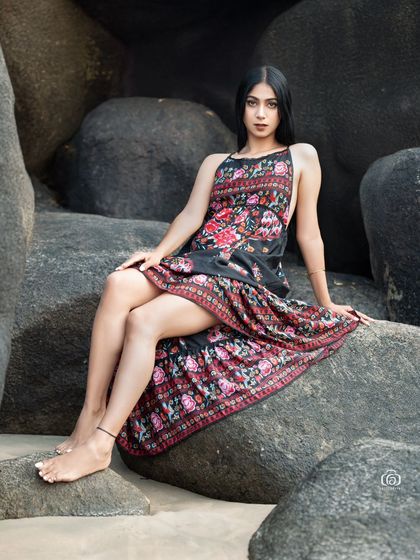 A different angle of the model in the floral dress, sitting on a large rock. The composition highlights the pattern of the dress against the neutral tones of the sand and rocks.
