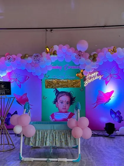 This butterfly naming ceremony decoration features a custom backdrop with the baby's photo. The organic balloon arch and butterfly details are illuminated with colored spotlights to create a magical, glowing effect in the evening.
