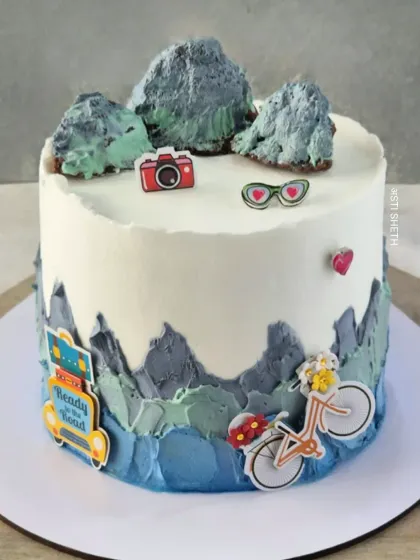 A surprise cake for a road trip lover, featuring mountains, a bicycle, and other travel-themed toppers on an Alphonso Vanilla cake.