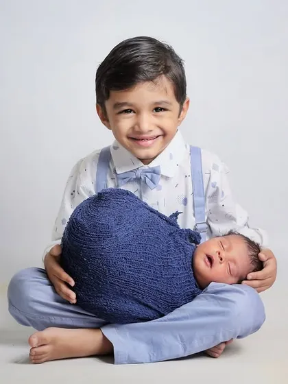 A proud big brother holds his sleeping newborn sibling on his lap. The coordinated blue outfits tie the whole image together beautifully.