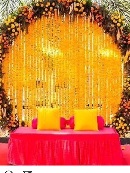 A vibrant outdoor Haldi photo booth with a circular backdrop of marigold strings. The bright pink seating provides a beautiful pop of color against the yellow flowers.