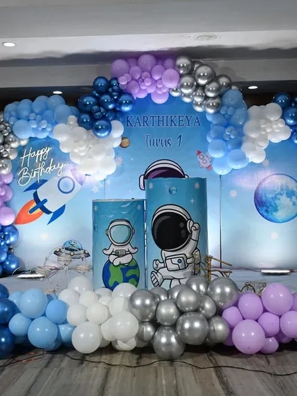 An out-of-this-world space and astronaut theme for a first birthday. This large stage setup features a galaxy backdrop, planet and rocket details, and a full balloon arch in shades of blue, purple, and chrome silver to create a cosmic adventure.