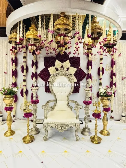 A beautiful setup for a seemantham or baby shower. The decor features a white throne surrounded by hanging floral arrangements and traditional temple bells, creating a divine atmosphere.
