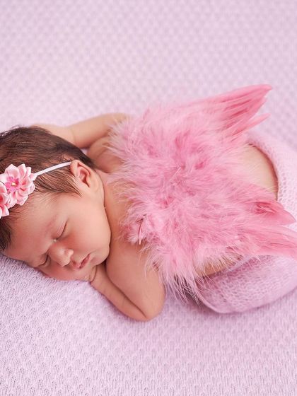 A tiny angel, this newborn is swaddled in pink with matching feathered wings and a floral headband, sleeping on a soft pink blanket.