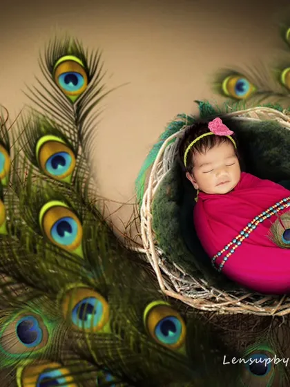 This newborn girl is styled as a divine little Radha, nestled in a basket adorned with peacock feathers. The vibrant pink wrap and serene expression create a beautiful and graceful portrait.