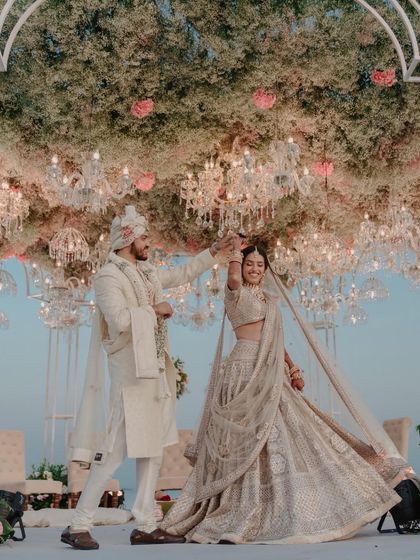 A playful twirl under a canopy of flowers and chandeliers. This action shot captures the bride's flowing lehenga and the couple's joyful interaction during their beachside wedding.