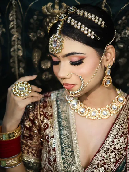 A classic bridal pose featuring a deep red velvet lehenga and kundan jewelry. The makeup is soft and timeless, with a focus on rosy cheeks and subtly defined eyes.