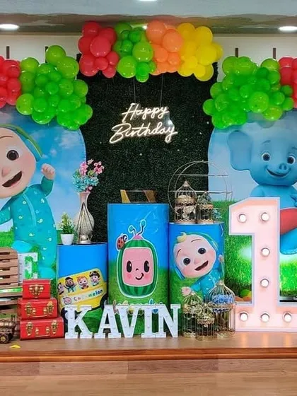 A bright and cheerful Cocomelon theme with a green hedge backdrop. The setup features character cutouts, a marquee number '1', and themed props like the Cocomelon bus.