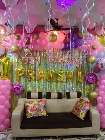 A first birthday party with a full pink and gold balloon setup. The decoration includes a dense balloon arch over the sofa, a shimmer curtain, and themed foil balloons on pillars.