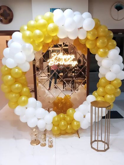 A simple and elegant birthday decoration with a circular balloon arch in gold and white. The backdrop features a mirrored panel that reflects the 'Happy Birthday' neon sign, adding depth to the setup.
