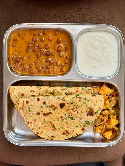 A plate with stuffed paratha, kala channa curry, sautéed potatoes, and curd. This is a hearty and filling meal, perfect for a weekend lunch.