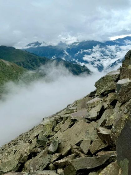 A path carved through a field of boulders, with clouds resting in the valley below. This trek is a journey through diverse and dramatic terrains.