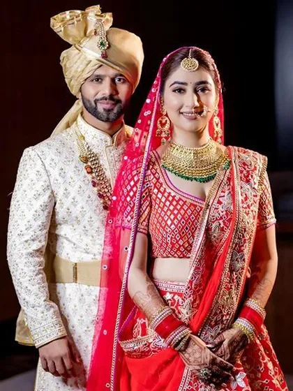 A beautiful example of a coordinated red and cream wedding outfit. The bride's red lehenga features a unique geometric pattern on the blouse, complemented by the groom's simple yet elegant cream sherwani.