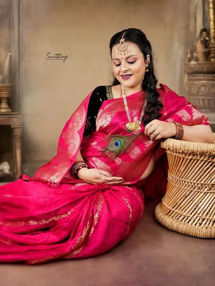 A serene portrait of a mother-to-be in a traditional pink saree, holding a peacock feather. The setting, inspired by classic Indian art, creates a rich and soulful atmosphere.