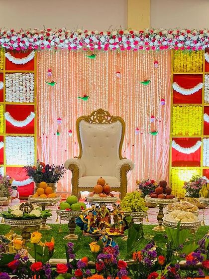 A colorful and festive Seemantham stage. The backdrop features red, yellow, and white panels with hanging garlands, and the foreground is filled with a beautiful array of flowers and offerings.