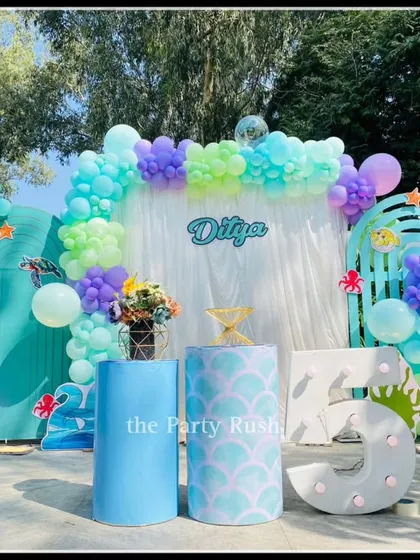 I create beautiful underwater worlds, like this mermaid-themed party for Ditya's 5th birthday. This outdoor setup uses a palette of seafoam green, purple, and blue, with shell cutouts, a shimmering backdrop, and a large marquee number '5' to make a splash.