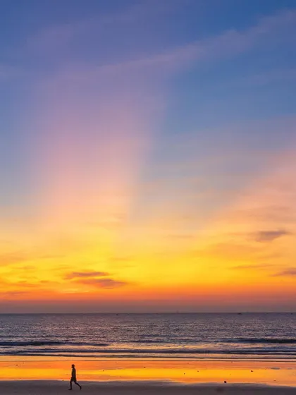 A lone walker on the beach during a spectacular winter sunset. The sky is filled with vibrant yellows, oranges, and purples, creating a breathtaking scene.