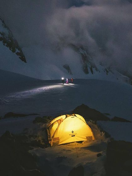 A glowing tent provides a beacon of warmth and safety in the vast, dark expanse of a snowy mountain pass. In the distance, fellow campers make their way with headlamps.