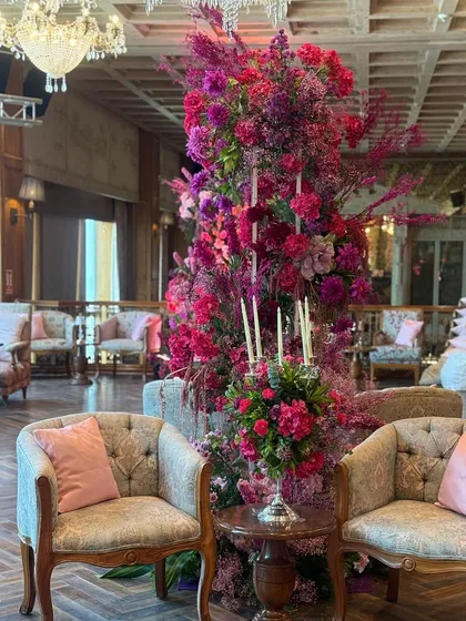 A tall, dramatic floral arrangement in shades of pink and magenta stands as a piece of art within the Mehendi celebration. Placed between elegant lounge chairs, it adds height and a bold splash of color to the space.