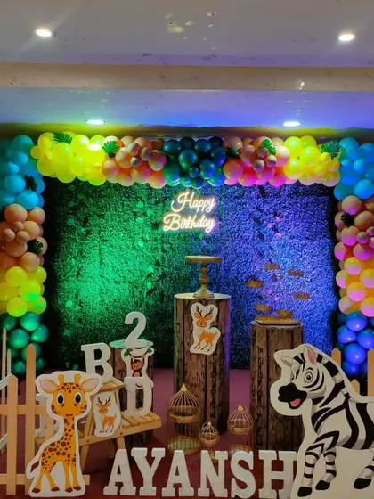 A colorful jungle party setup with a vibrant, multi-colored balloon garland over a grass backdrop. The scene is lit with fun colors and features cute animal props.