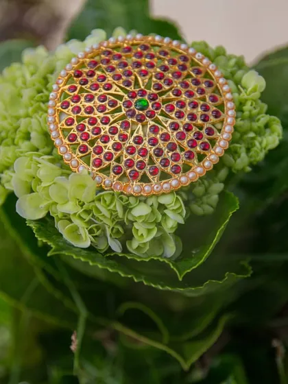A detailed photograph of a 'jadai billai', a traditional hair ornament used in South Indian weddings. We focus on capturing the intricate red and green stonework and pearl details.