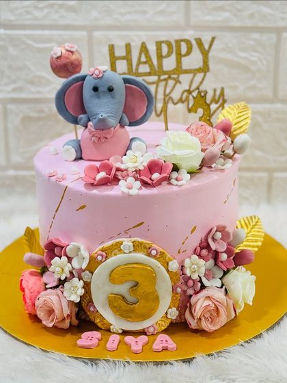 A pretty-in-pink elephant cake for a third birthday. This sweet design is adorned with a cute fondant elephant, beautiful flowers, and golden accents.
