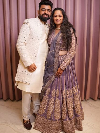 A happy couple posing together. She wears the mauve-purple lehenga, and he complements her in a classic white sherwani.