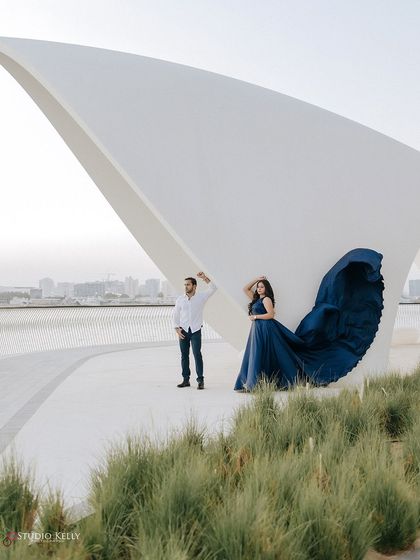 A dramatic portrait in Dubai, where the bride's flowing blue gown contrasts beautifully with the clean white lines of the modern architecture. The wind catching the fabric creates a stunning visual effect.