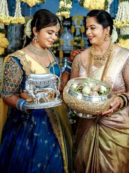 Two women at a traditional ceremony, showcasing coordinated blue and gold ensembles. Their outfits, while different in style, share a common color palette and level of intricate detail, creating a unified look.
