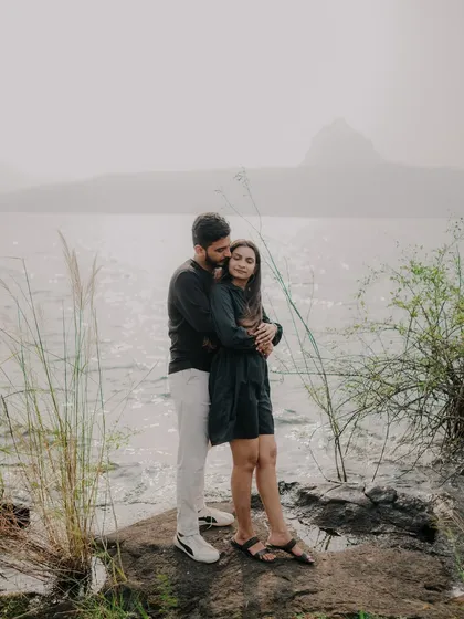 A wide shot of the couple embracing on the rocks by the lake. The misty mountains in the background add a sense of grandeur and drama to this romantic scene.