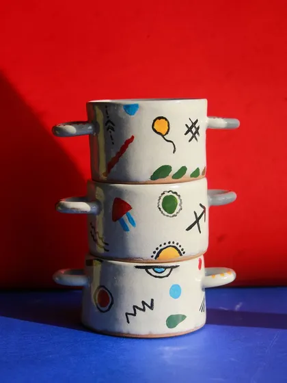 A stack of small, two handled bowls from the 'Playful Mornings' collection, decorated with colorful, abstract symbols against a bright red background.